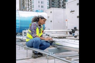 Woman in safety gear working on a laptop near a large factory air conditioner