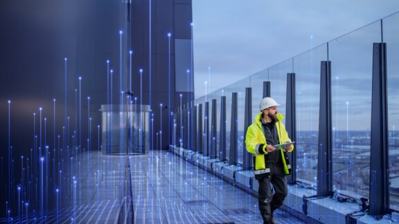 Man in high visibility jacket and safety helmet walking along the corridor of an office building with a tablet in hand
