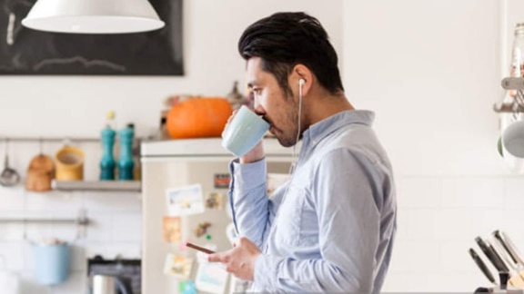 A businessman sipping on tea in a kitchen wearing wired headphones