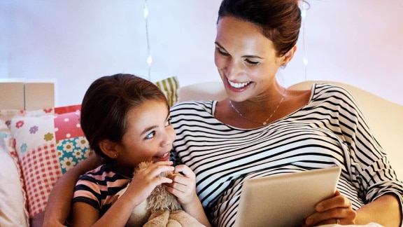 A smiling mother and child in bed chatting and looking at a tablet