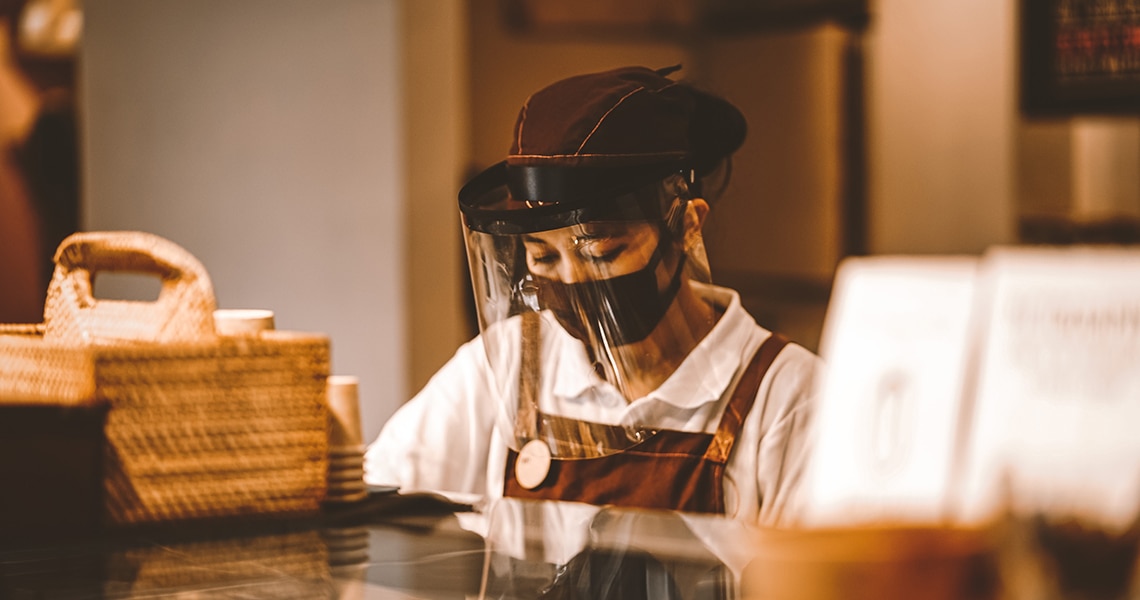 A barista wearing a face-mask and face-shield, working behind a counter