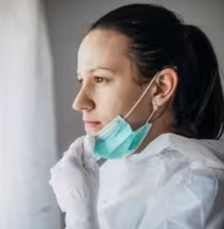 A female emergency worker gazing out a window while removing her face-mask