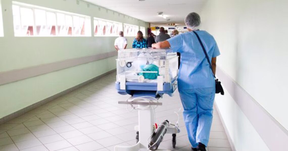 Rear view of a medical professional in scrubs wheeling a stretcher along a passage, with people walking ahead