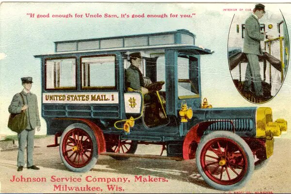 A man driving a 1911 United States Mail Truck with another man standing by it with a bag