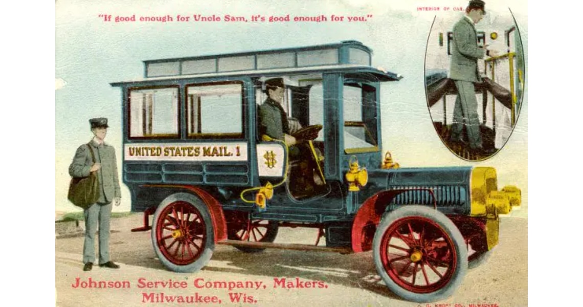 A man driving a 1911 United States Mail Truck with another man standing by it with a bag