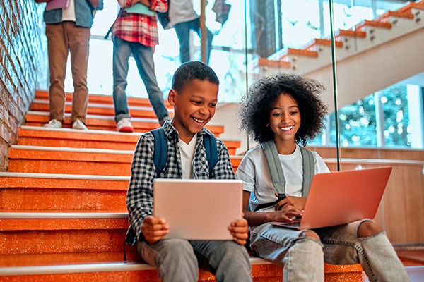 Two children sitting on the stairs and smiling while working on a laptop